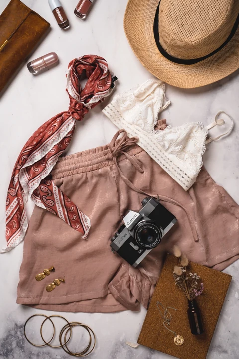 flat lay of a white lace top, pink shorts, a straw hat, red scarf, and accessories